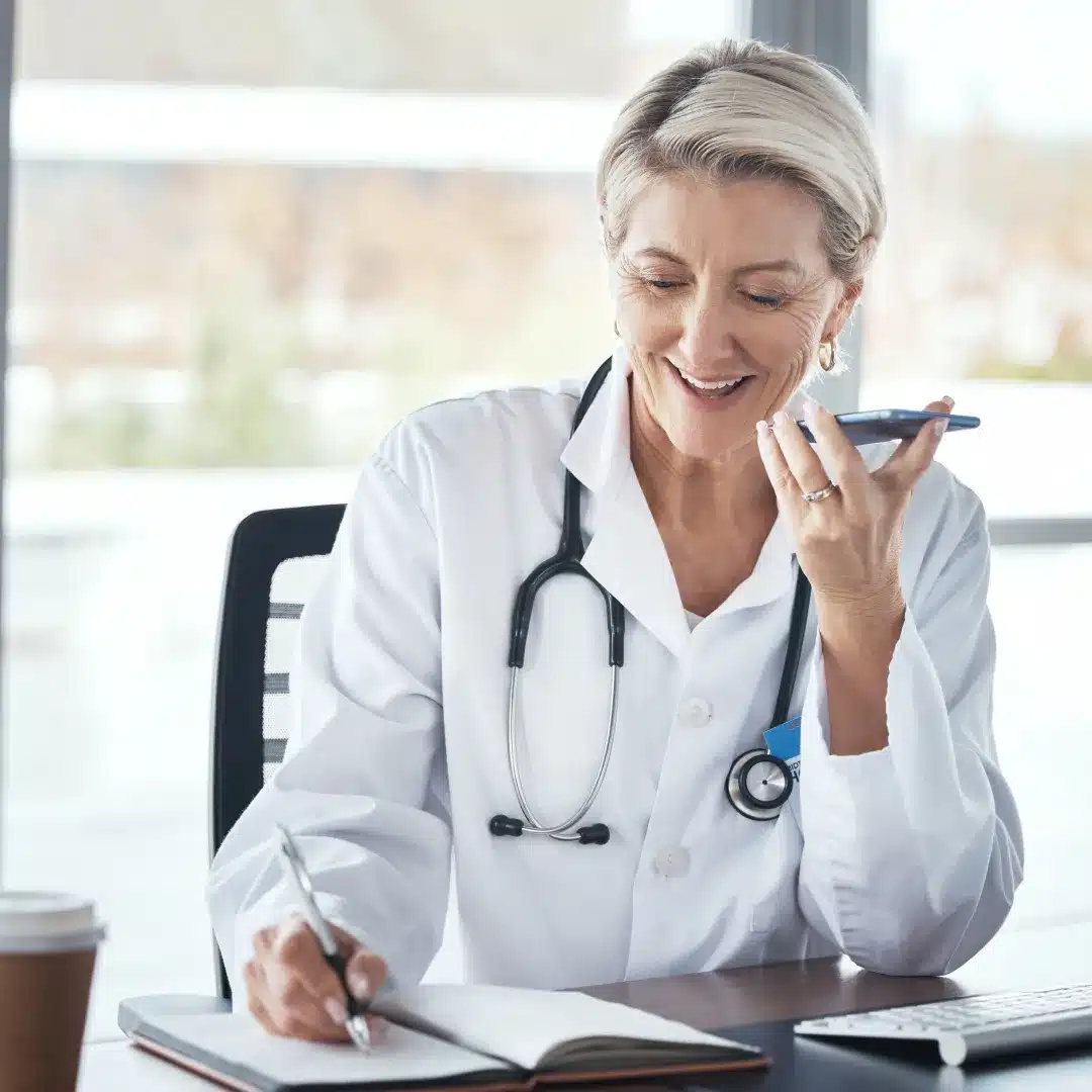 Senior female physician in a white coat speaking on a phone while taking notes at her office desk.