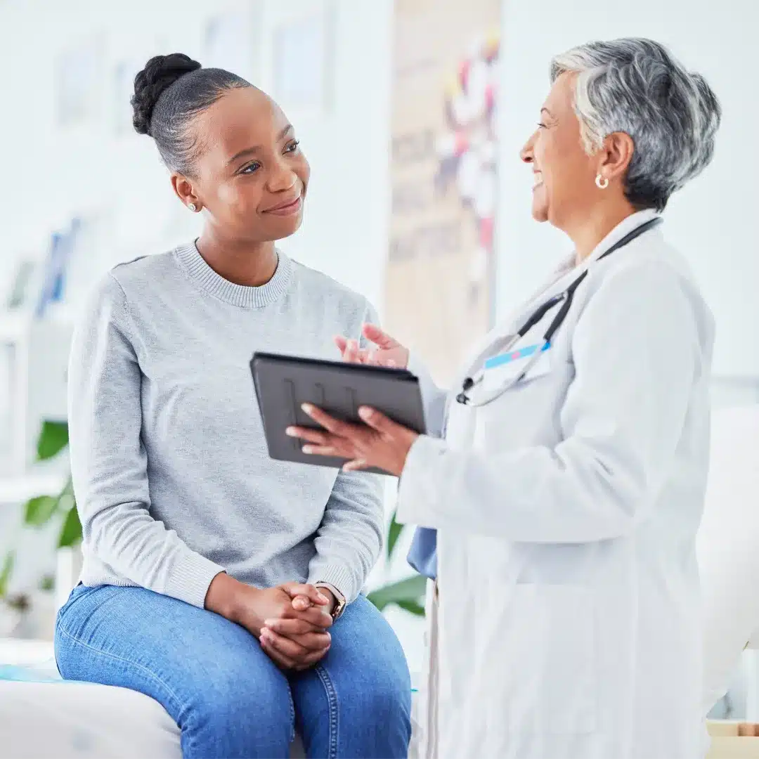 Senior female physician with a stethoscope reviewing a tablet with a patient in a bright exam room.