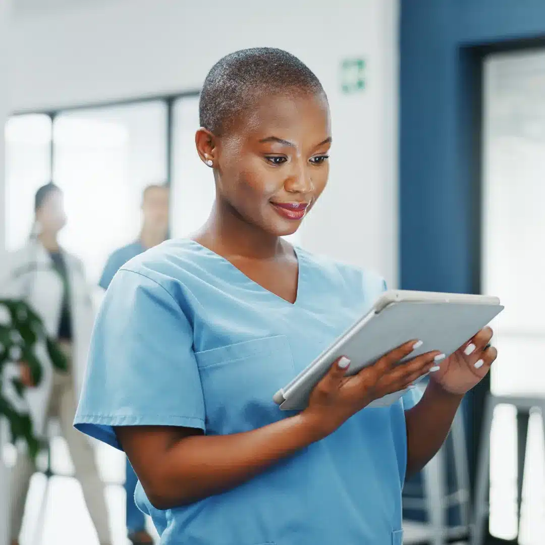 Clinical staff member in light blue scrubs and stethoscope reviewing a tablet in a hospital hallway.