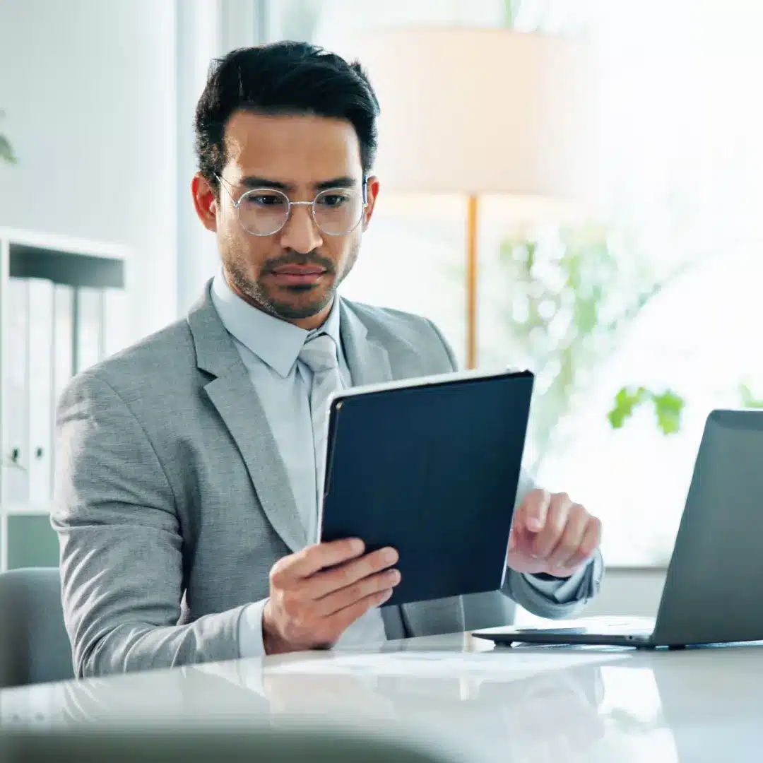 Young man in a gray blazer and glasses reviewing a tablet at a desk with a laptop open beside him.