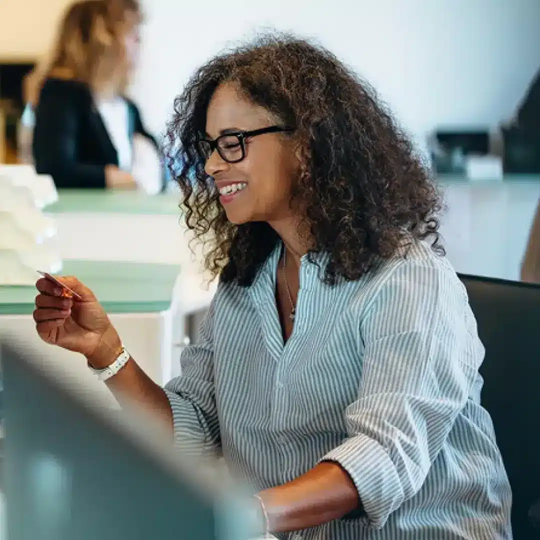 Smiling woman with curly hair and glasses holding a pen at an office desk with a monitor in view.