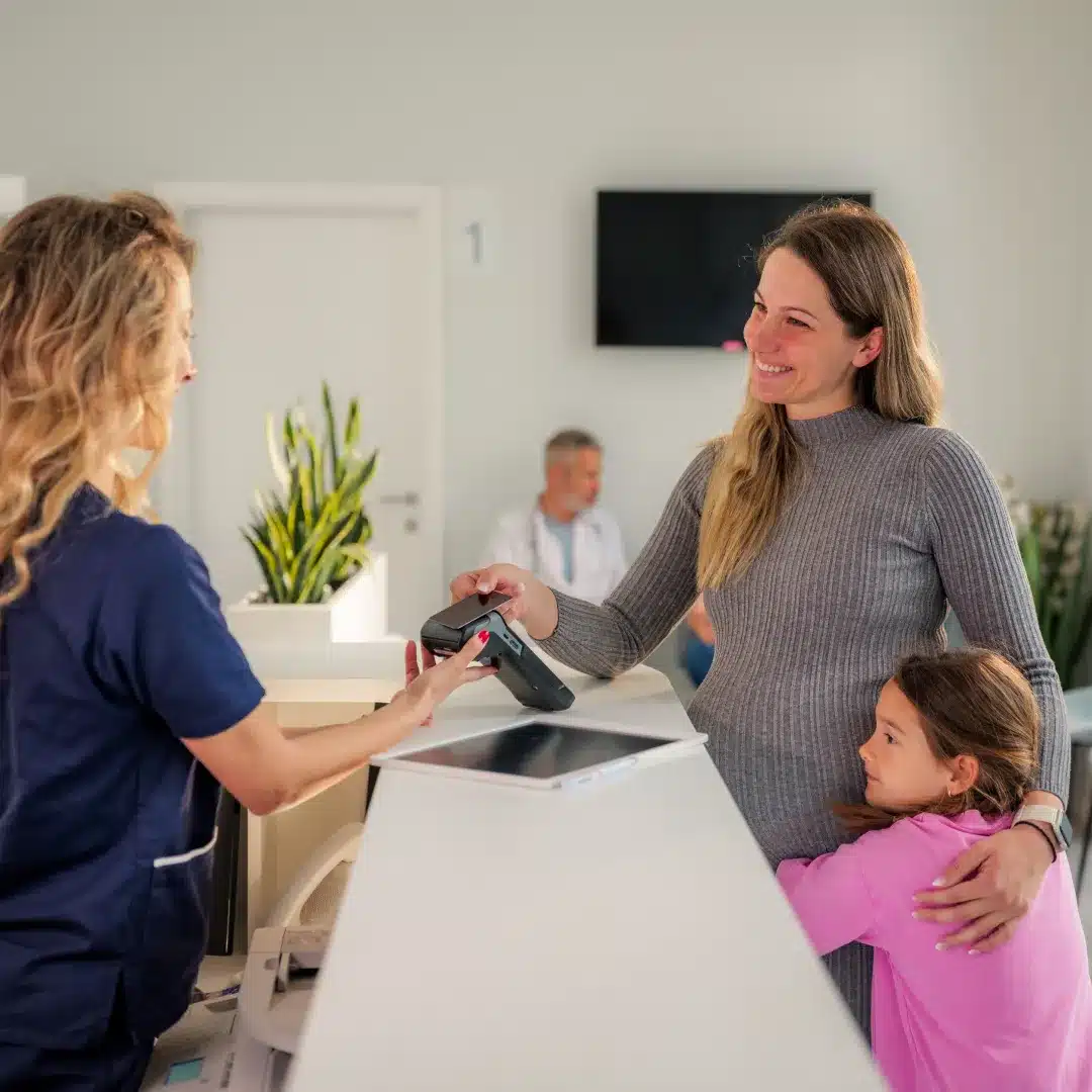 Patient and child checking in at a medical front desk while a nurse processes a card payment.