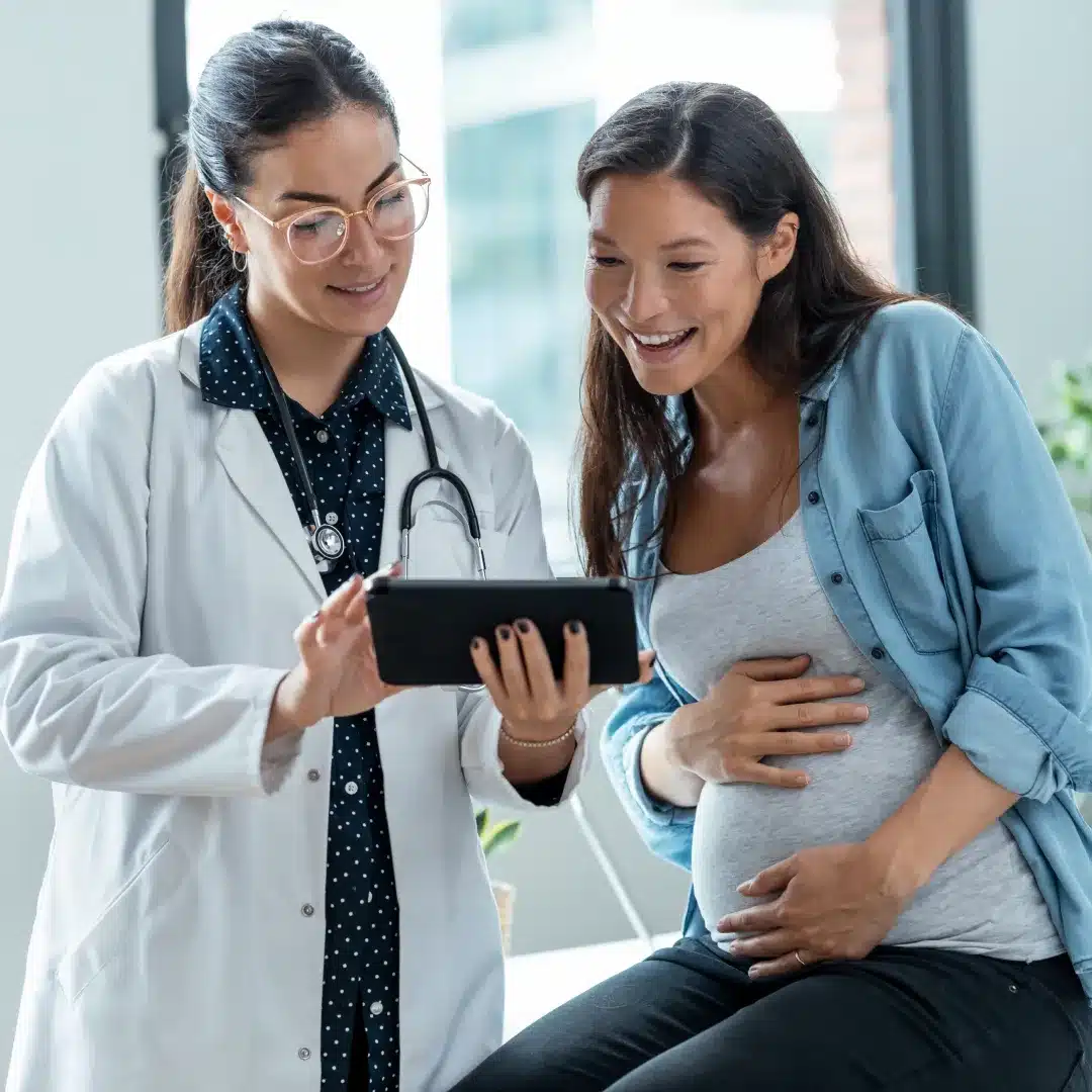 Female physician in a white coat showing a tablet to a smiling pregnant patient during a clinic visit.