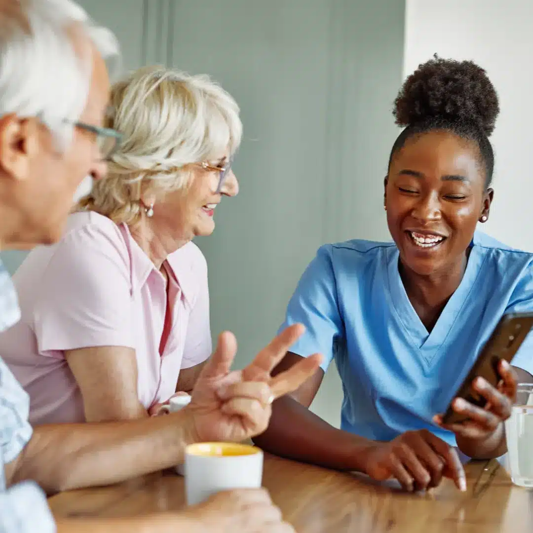 Nurse in light blue scrubs showing a smartphone to two elderly patients at a table in a care setting.