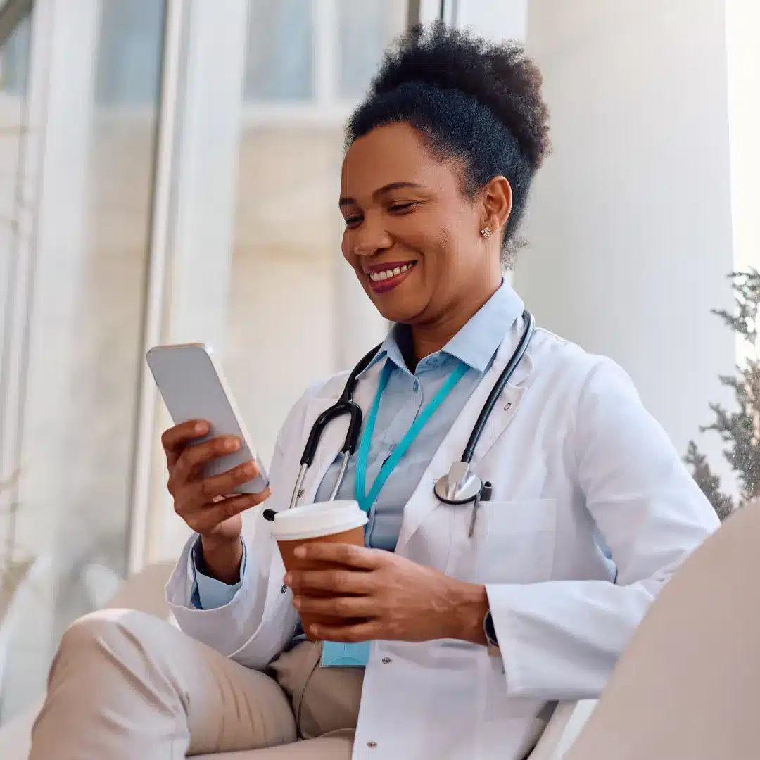Female physician in a white coat and stethoscope checking her phone while holding a coffee cup.