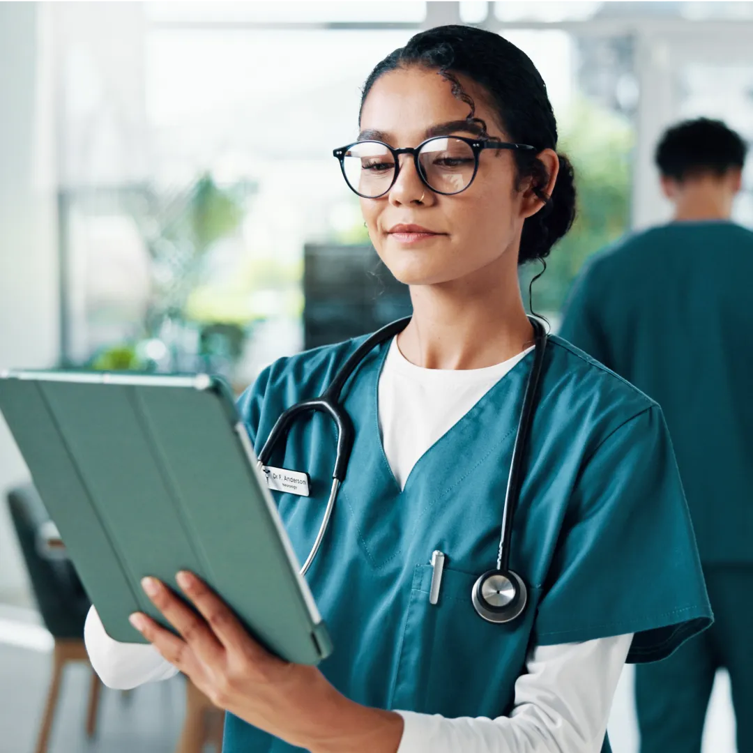 Clinical staff member in teal scrubs and glasses reviewing a tablet in a medical facility hallway.