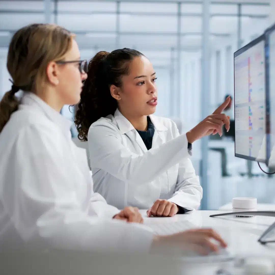 Two women in white lab coats are collaborating at a desk, discussing data displayed on a computer screen.