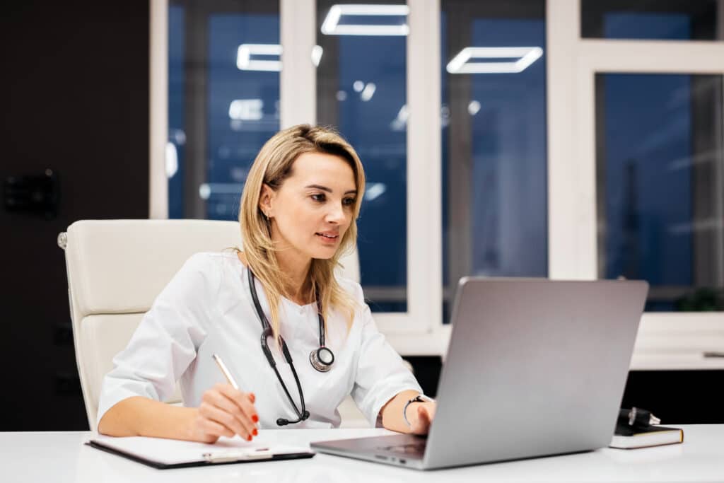 Female doctor with stethoscope is focused on her laptop at a modern office desk, notepad and pen beside her, large windows in the background